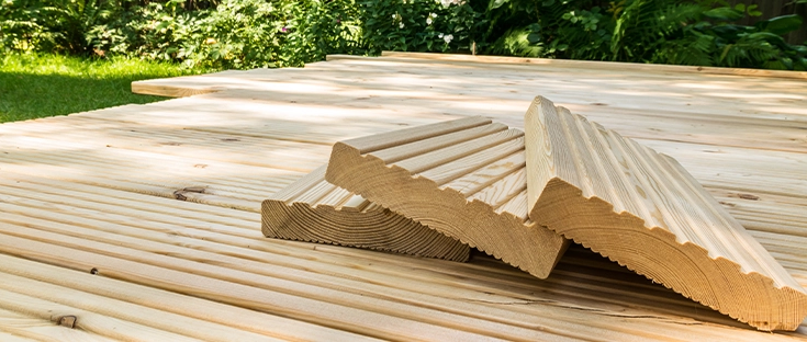 Terrassenbau mit hellen Lärchenholzdielen, gestapelte Riffelbretter im Vordergrund, umgeben von grünem Garten.