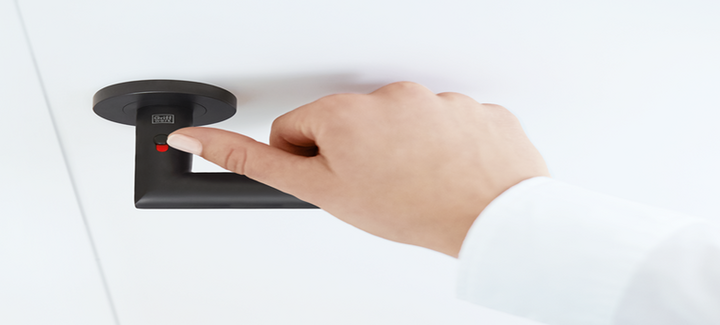 Hand bedient den roten Fingerprint-Sensor des schwarzen Türgriffs me time mit Smart2Lock-Technologie an einer weißen Tür.