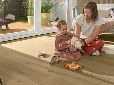 Helles Parkett im Wohnzimmer. Eine Frau spielt mit einem kleinen Mädchen auf dem Boden.